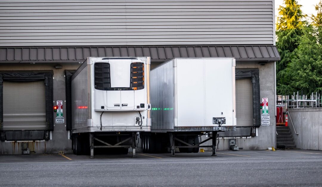 Two trailers backed into warehouse loading docks beside a metal-sided building, with pavement and trees nearby.