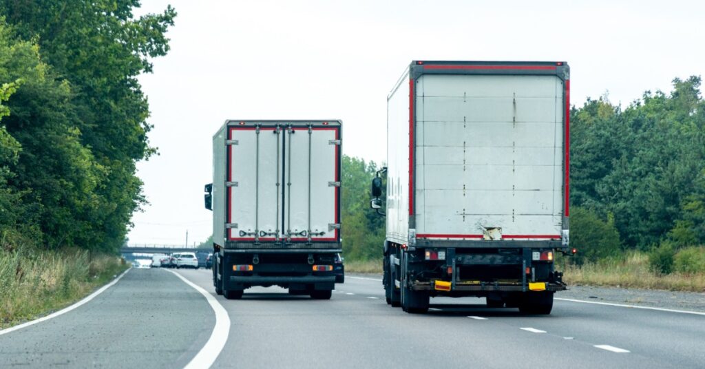 Two box trucks traveling on a tree-lined highway, with several cars and an overpass visible in the distance.