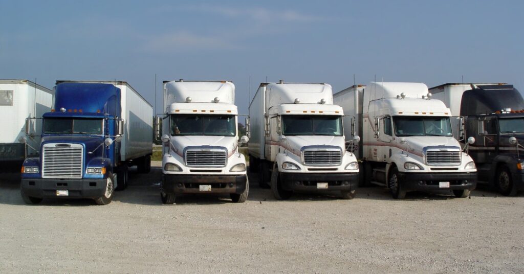 Several semi trucks parked side by side on a gravel lot under a clear blue sky, including blue, white, and black cabs.