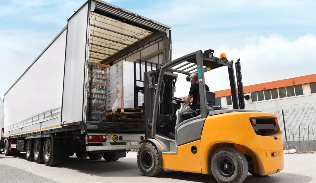 A person drives a yellow forklift loading stacked pallets into an open trailer outside a warehouse under a cloudy sky.