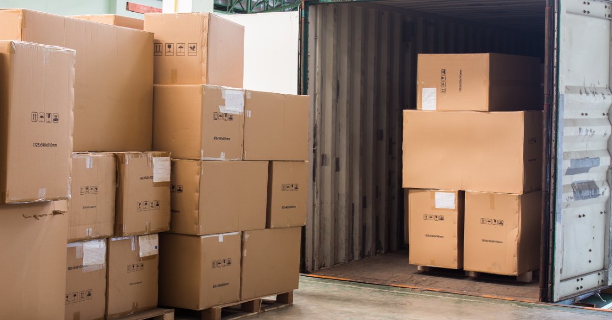Large cardboard boxes stacked on wooden pallets beside an open shipping container holding more boxes inside a warehouse.